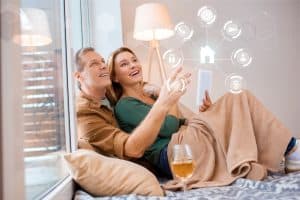 smiling wife holding digital tablet while sitting floor wth husband adjusting the smart home