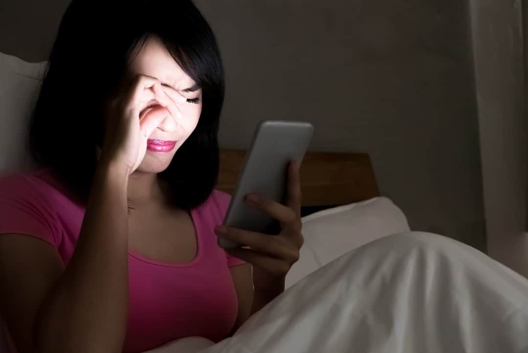 Woman on a bed in the dark staring sadly at a cell phone.