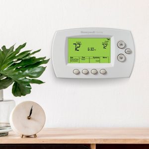 Honeywell RTH6580WF 7 day thermostat on a wall. Shelf with a plant and clock in the foreground.
