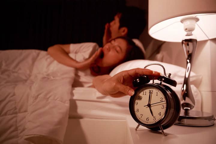 Alarm clock and lamp in foreground, young couple in bed in background.