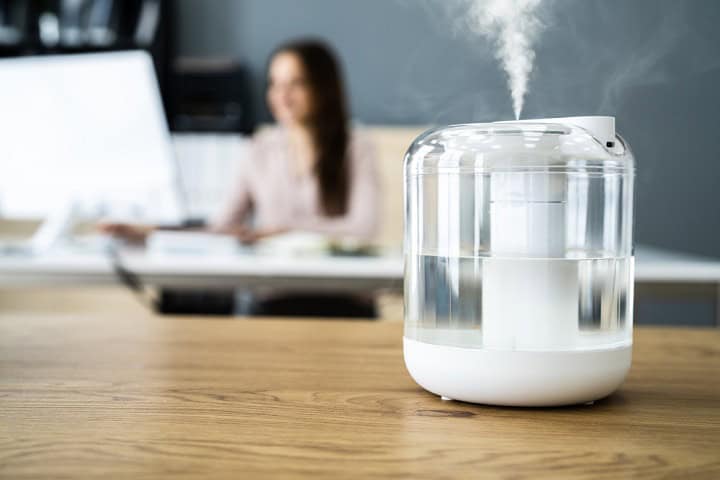 Humidifier diffusing mist into the air with a woman working at a desk in the background.