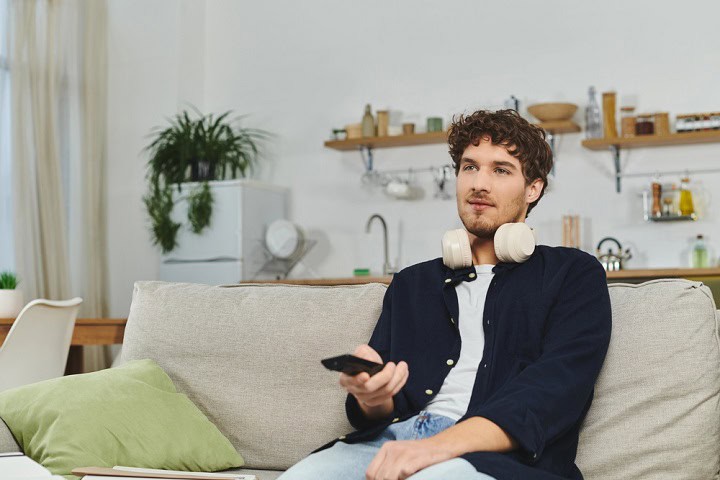 Man on a couch holding a remote and wearing headphones.