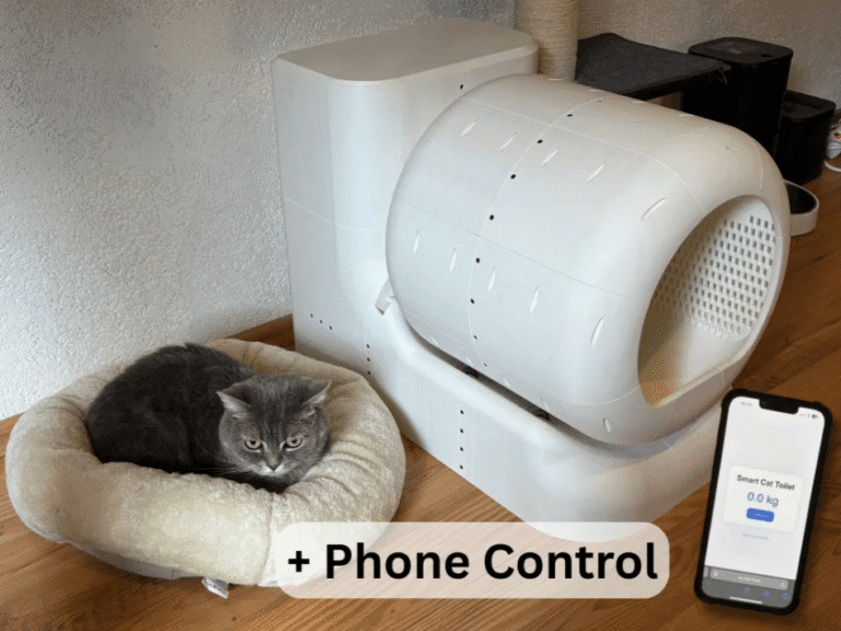 Cat in a cat bed sitting next to the SmartCat Toilet. Cell phone in the foreground showing the interface for the litter box.