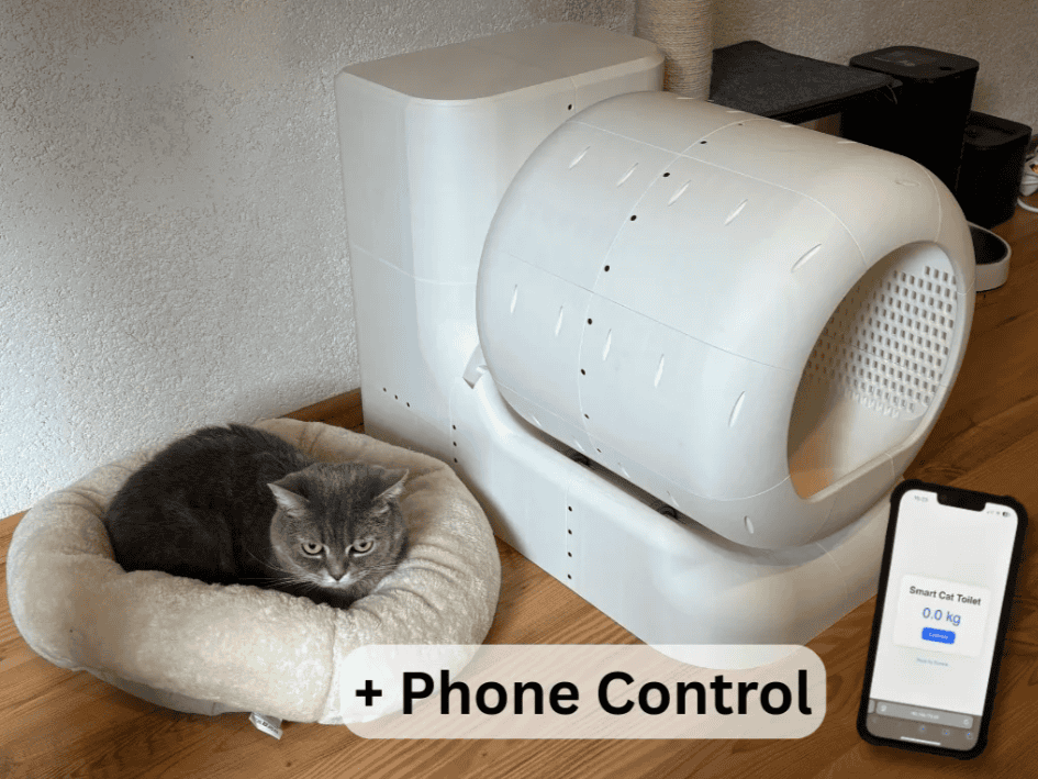Cat in a cat bed sitting next to the SmartCat Toilet. Cell phone in the foreground showing the interface for the litter box.