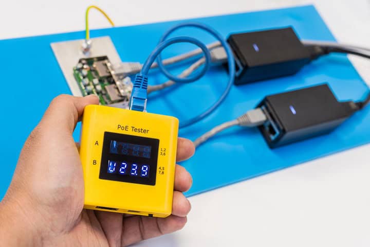Power over ethernet tester in foreground with networking setup in background.
