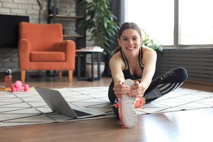 Woman stretching on the floor with a laptop sitting beside her.