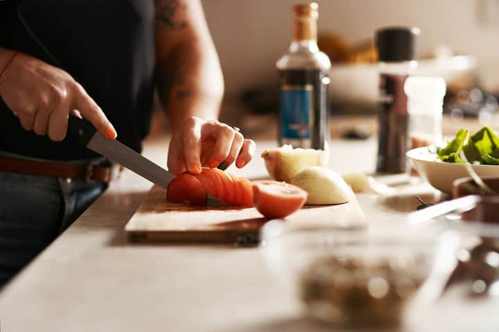 Man cutting a tomato on a cutting board, cooking utensils in foreground and background.