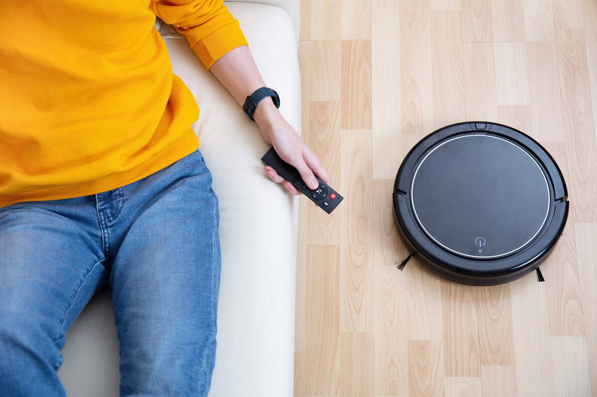 View of a person laying on couch with a remote, while a robot cleaner runs below them cleaning the floor.